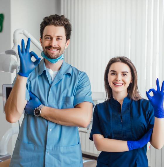 Photo of smiling dentist standing with arms crossed with her colleague, showing okay sign.