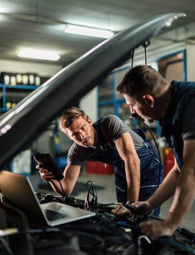 young-car-repairman-talking-with-his-coworker-while-checking-engine-performance-with-diagnostic-tools-auto-repair-shop